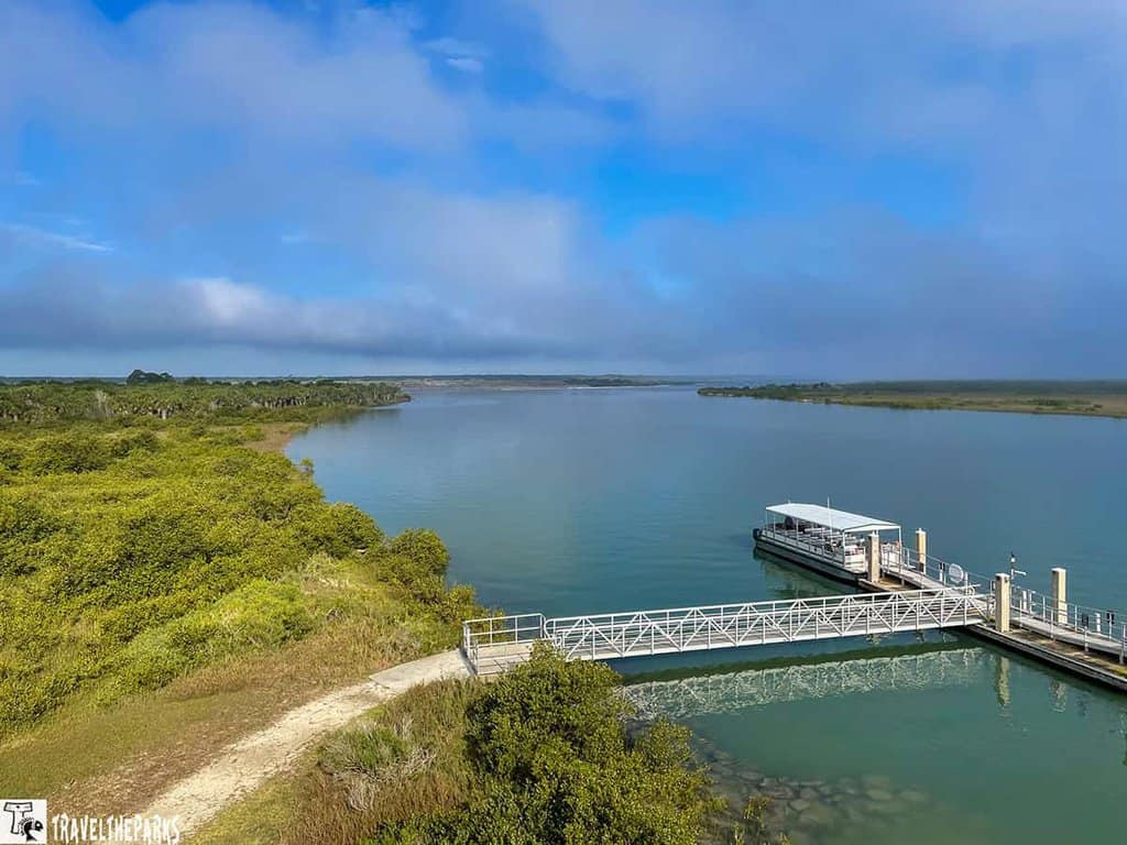 an expansive view of the Matanzas river surrounded by lush greenery under a vibrant blue sky partially covered with clouds. In the foreground, there is a white dock extending into the water with a small covered ferry boat moored beside it. The water appears calm and reflective, enhancing the tranquil atmosphere. The surrounding landscape consists of dense green vegetation on the left and stretches into the distance, highlighting the serene natural setting.
