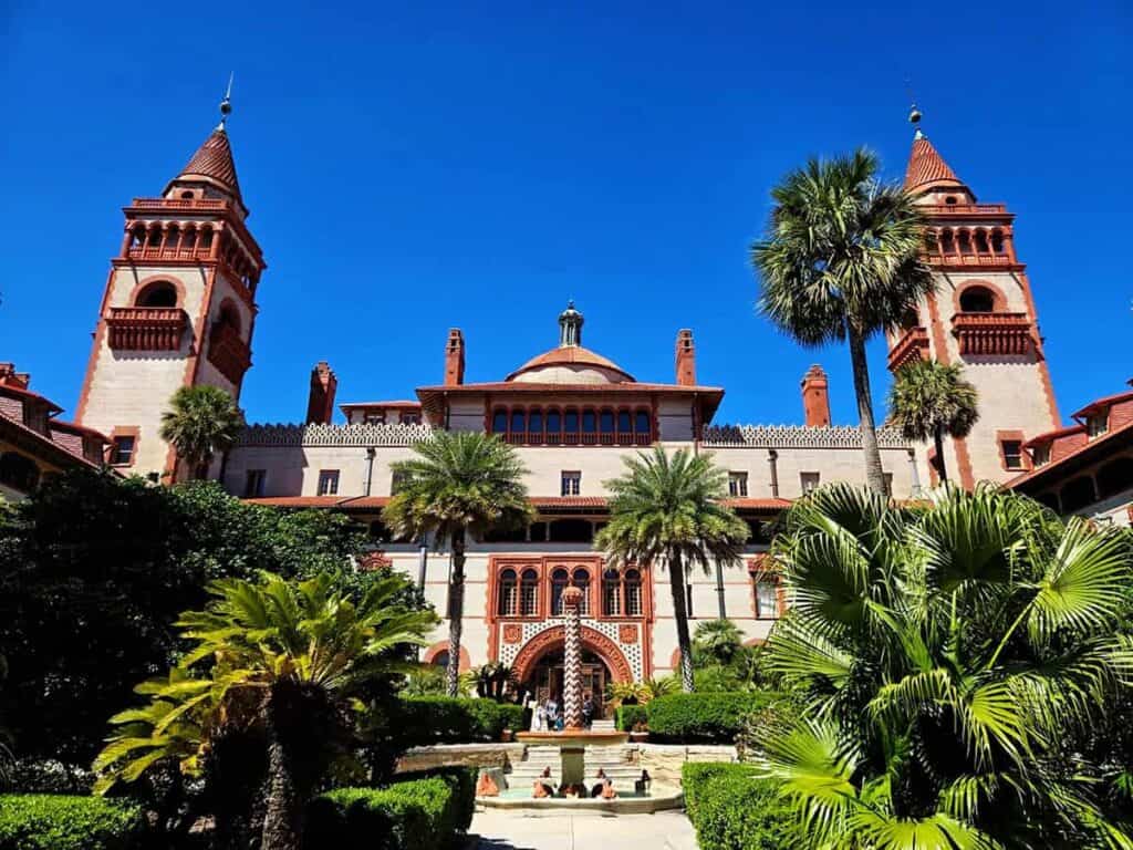 the facade of Flagler College in St. Augustine, showcasing a grand and historic architectural style. The building features two prominent red-brick towers with pointed turrets on either side, reaching high into a clear blue sky. The central structure has a large, arched entryway with intricate designs, flanked by symmetrical rows of tall windows. In front of the building is a well-manicured garden with lush green palm trees and various shrubs. A stone pathway leads up to the entrance, and a small fountain is visible in the foreground, adding to the serene and majestic atmosphere.