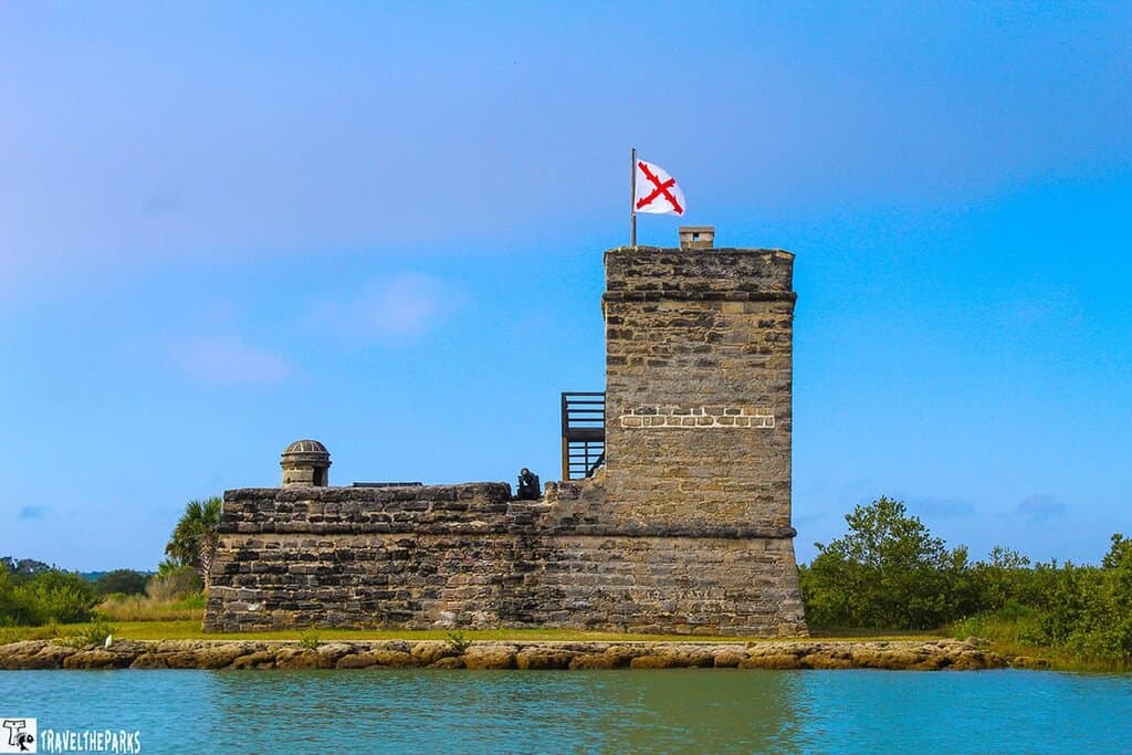 Fort Matanzas, a historic fortification made of coquina stone, located on the edge of a body of water. The fort has a square tower, with a flag flying at the top. The flag is white with a red cross. The tower is fitted with narrow openings and features two visible levels. To the left of the tower, a lower section of the fort displays similar stonework and includes a small dome-like structure. Surrounding the fort are lush green trees and shrubs. The sky above is bright blue with scattered clouds, and the water in the foreground is a clear turquoise, gently lapping against a stone shoreline.
