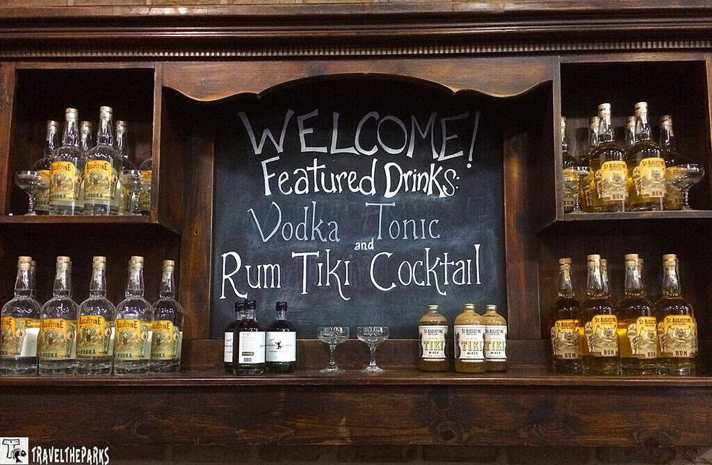 a wooden bar display with shelves holding various bottles of alcohol. In the center, there's a chalkboard with the welcoming message "WELCOME! Featured Drinks: Vodka Tonic and Rum Tiki Cocktail" written in a casual, handwritten style. The shelves flanking the chalkboard contain several clear bottles with light-colored labels, arranged neatly in two rows on either side. Below the chalkboard, there are two labeled bottles and two empty glass goblets. The wood has a rich, dark brown finish, giving the display a rustic appearance.