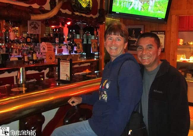 Two people smiling in a Western-themed Million dollar Cowboy Bar in Jackson with a TV showing a landscape.