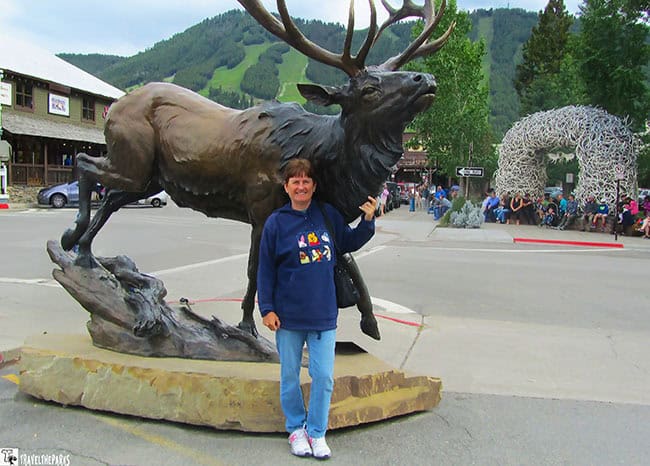 A person stands in front of a large elk sculpture on a stone base with a stop sign and an antler archway in the background.