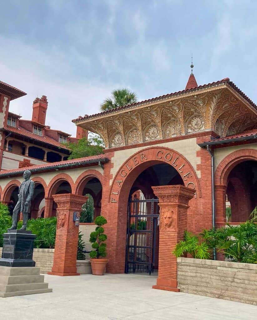 the entrance to Flagler College, showcasing a grand architectural structure with red brickwork and ornate detailing. The entrance is marked by a series of arches supported by columns with decorative carvings, including a lion's head. Above the central arch, gold lettering spells "FLAGLER COLLEGE." Overhead, elaborate designs cover the architecture, and a palm tree is visible, adding greenery to the scene. In the foreground, a statue of a person stands on a stone pedestal, adjacent to manicured shrubs in planters. The sky appears slightly cloudy.