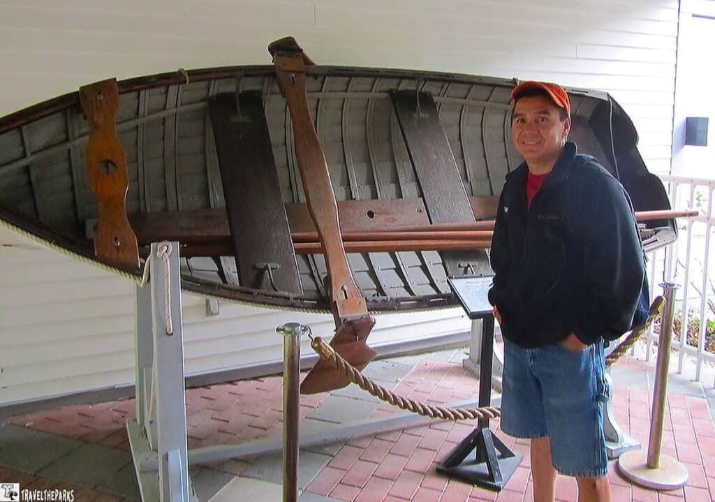 a man standing beside a vintage wooden boat displayed vertically on a white platform. The boat is secured with metal supports and positioned next to a white wall, showcasing its interior structure. The wooden surface of the boat is weathered, displaying rich brown hues. The man is wearing a red cap, a dark jacket, and denim shorts, with his hands in his pockets. A rope barrier in front separates the display from the walkway, which is laid with red and gray square tiles. A small informational plaque is visible near the base of the boat, giving context to the exhibit.