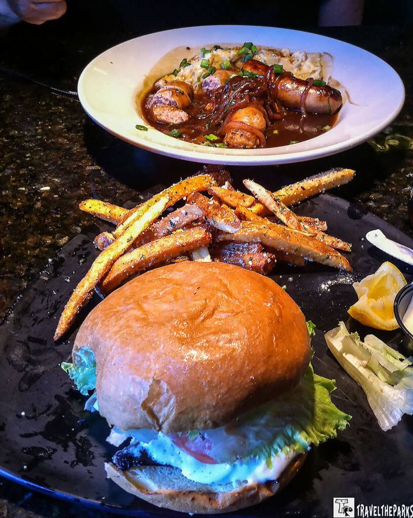 Delicious Meehan's Irish Pub: a plate of food on a dark, glossy table surface. In the foreground is a black plate with a burger and a side of fries. The burger features a glossy bun with a crisp lettuce leaf and a hint of sauce visible. The fries are golden, with a textured, crispy appearance and sprinkled with seasoning. To the side, there are light-colored vegetable garnishes along with a lemon wedge and a small dish of sauce. In the background, a white bowl contains sausages in a rich brown gravy, topped with green herbs and served over a bed of risotto.