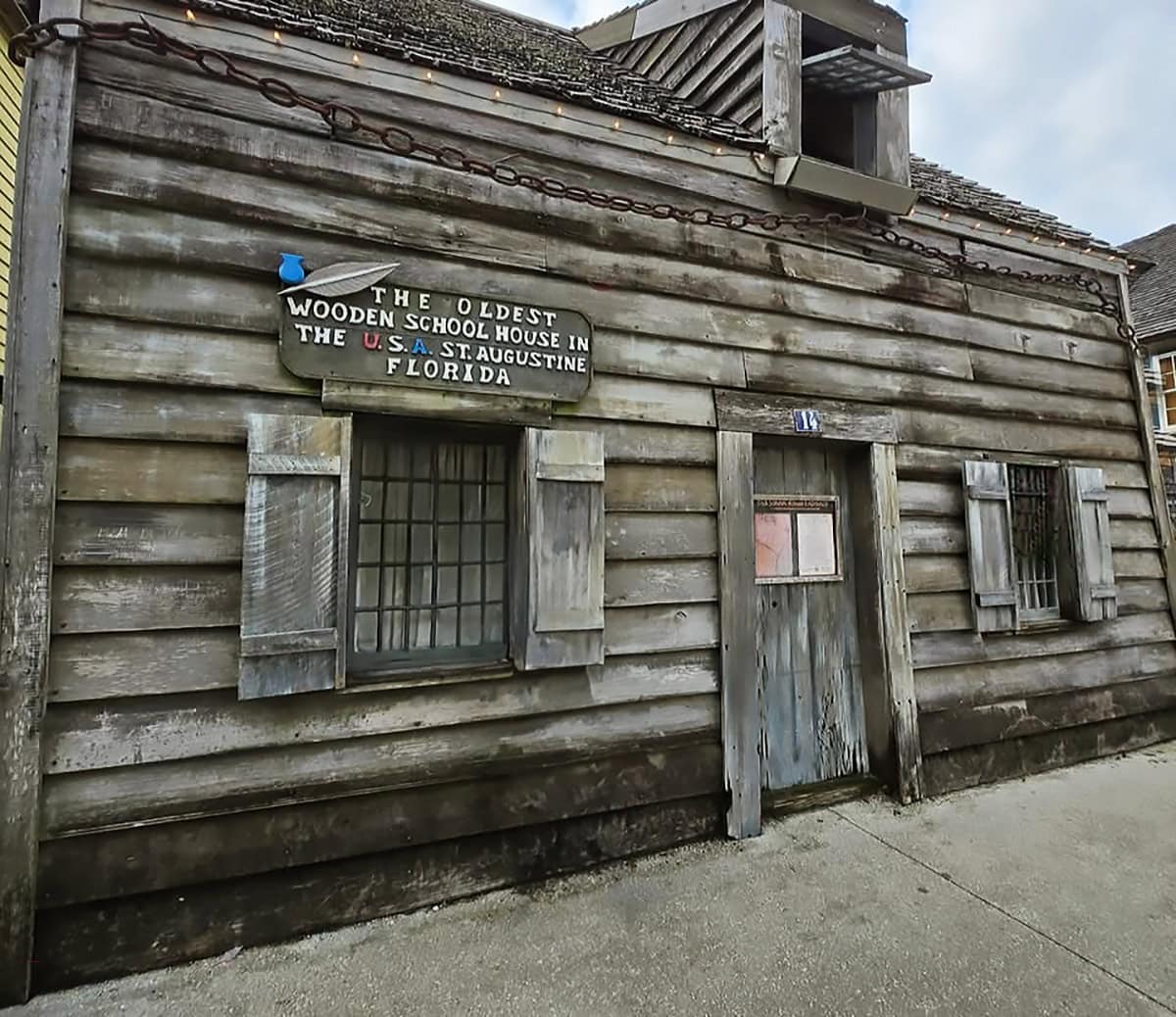 an old wooden building, identified as the oldest wooden schoolhouse in the U.S.A., located in St. Augustine, Florida. The structure is composed of aged, weathered wooden planks with visible horizontal lines. The front of the building features two windows, each with wooden shutters, flanking a central wooden door. Above the door is a small, rectangular sign with white, red, and blue lettering. A chain runs along the perimeter of the roof, contrasting with the muted tones of the weathered wood. The building rests on a concrete surface, and the sky is overcast.