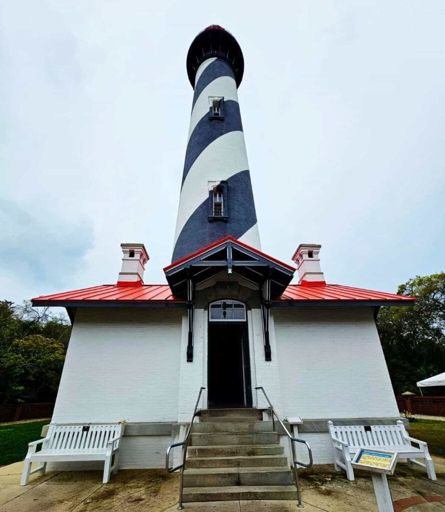 he St. Augustine Lighthouse, a tall cylindrical structure with a distinctive black and white spiral pattern. The lighthouse is prominently situated against a cloudy sky. The base of the lighthouse is a white, rectangular building with a red roof and two symmetrical chimneys on each side. A set of stone steps leads up to a black-framed entrance with a small overhang. Two white benches are placed symmetrically on either side of the entrance. A small sign is visible near one of the benches. Surrounding the lighthouse building, there is greenery and a partial view of a low brick wall.