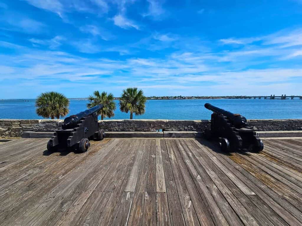 two black cannons mounted on wooden carriages situated on a wooden deck. The cannons are facing a large body of water under a bright blue sky with scattered white clouds. Behind the cannons, there is a low stone wall, and beyond it, three palm trees are visible on the left. Across the water, a distant view includes a bridge connecting land areas. The water is calm, reflecting the clear sky, adding a serene contrast to the historical artillery in the foreground.