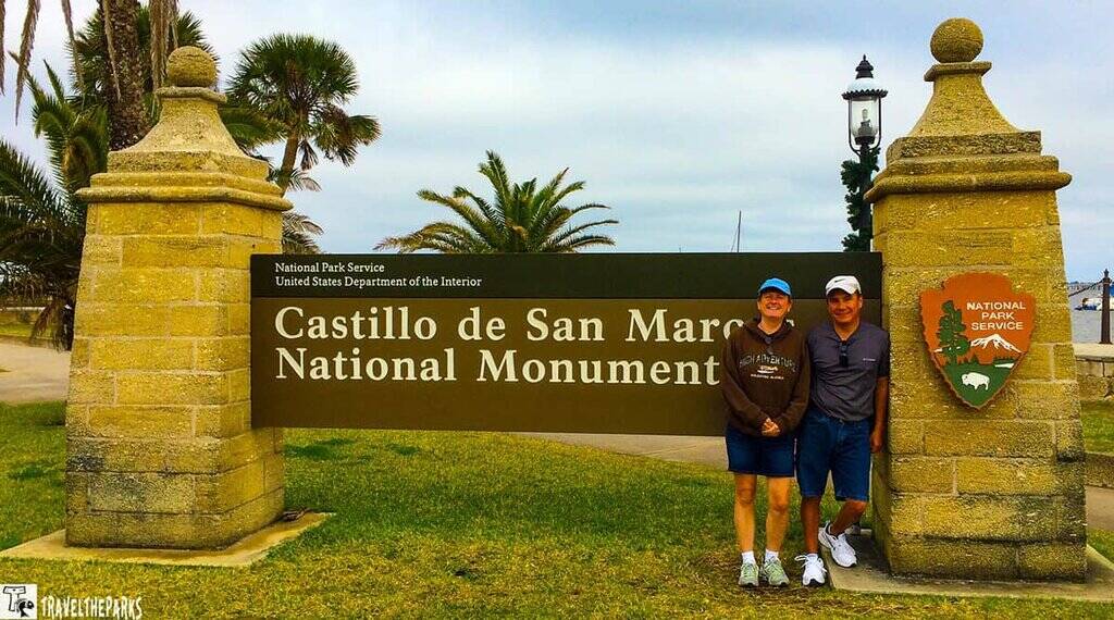 two people standing in front of the sign for Castillo de San Marcos National Monument. The sign is supported by two stone pillars with decorative tops. Behind the sign, there are tall palm trees, adding a tropical feel to the scene. The grass in the foreground is well-maintained, and the sky appears overcast. A body of water and a lamp post are visible in the background, contributing to the picturesque setting.