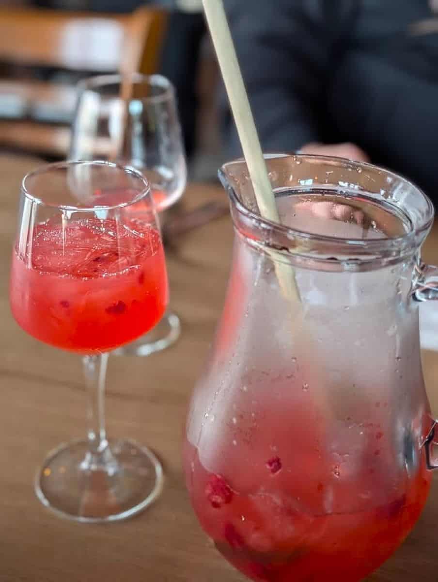 a close-up view of a dining table with a pitcher and a glass filled with a red beverage. The pitcher is large and made of transparent glass, containing ice cubes and red fruits, with a wooden stirrer placed inside. In the foreground, a stemmed glass also holds the same red drink. The wooden table surface is visible, and in the background, there are two blurred wine glasses, suggesting a dining or restaurant setting. The overall ambiance appears casual and relaxed.