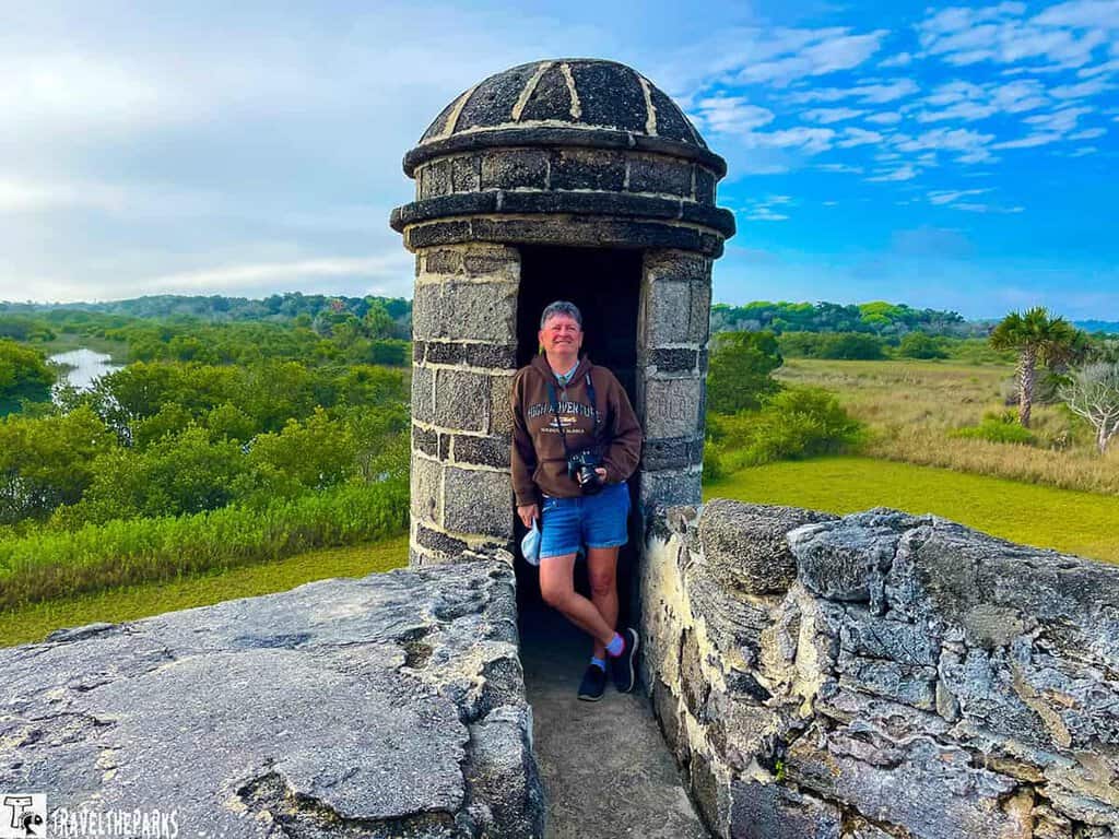 Fort Matanzas, featuring a small stone watchtower. A person stands inside the tower doorway, leaning casually against the stone structure. The tower's cylindrical shape is topped with a dome. The surrounding landscape is lush and green, with trees and open fields stretching to the horizon under a partly cloudy blue sky. The foreground showcases the textured stonewalls of the fort, exhibiting wear and natural aging.
