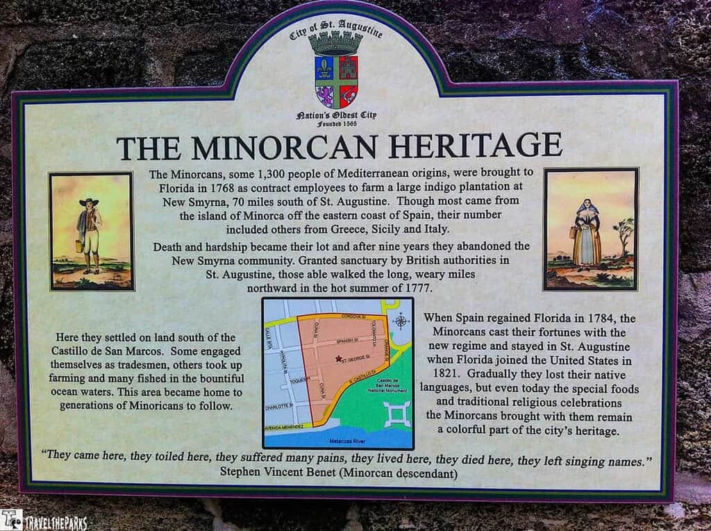 an informational sign titled "The Minorcan Heritage" set against a textured, dark stone wall. The sign features the City of St. Augustine's coat of arms at the top, alongside the text "Nation's Oldest City Founded 1565." The main text describes the history of the Minorcans, Mediterranean people brought to Florida in 1768. It details their journey, hardships, and eventual settlement in St. Augustine after leaving New Smyrna. There are two drawings on either side; the left depicts a man with a broad-brimmed hat, a cane, and a simple outfit. The right shows a woman in traditional attire holding a basket. A small colored map in the bottom center illustrates a part of St. Augustine, highlighting areas of importance. A quote by Stephen Vincent Benet, a Minorcan descendant, is at the bottom of the sign.