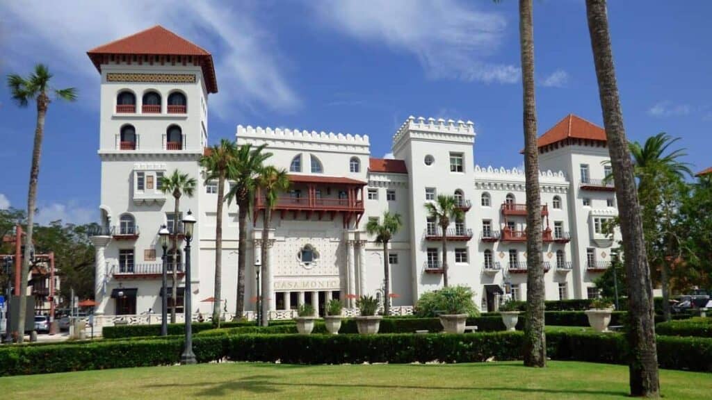 The Lightner Museum in St. Augustine, Florida. The structure is a large, historic building painted in white with multiple floors and intricate architectural details. The facade features arched windows, balconies with red railings, and a distinctive roof with red tiles. The building has a symmetrical design, with tall square towers on each end. Several palm trees are in the foreground, adding to the tropical setting. Well-maintained hedges and a neatly trimmed lawn lead up to the building.