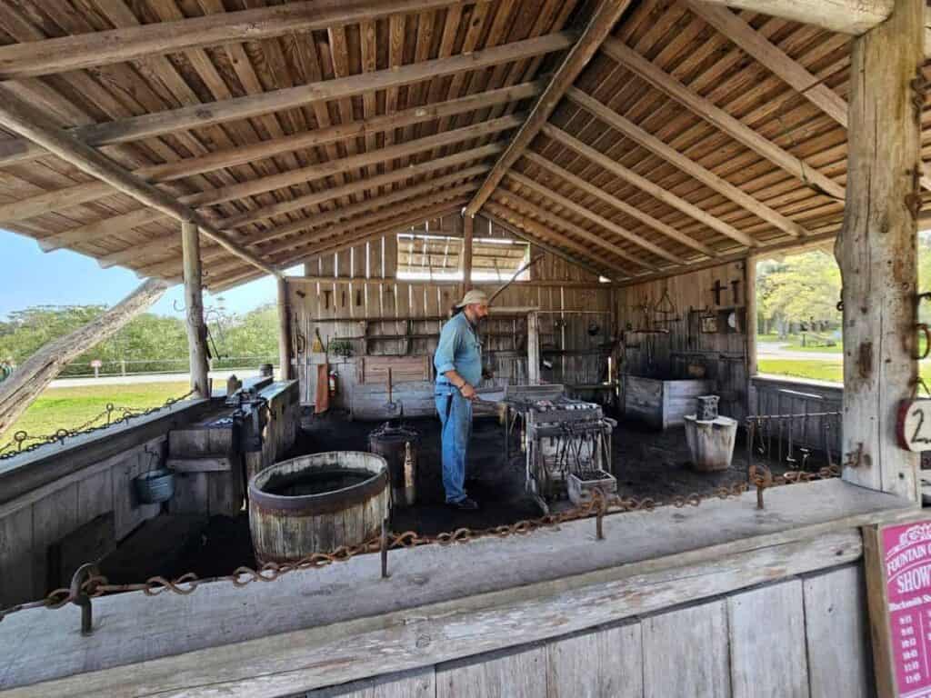 the interior of a rustic blacksmith workshop. A man wearing a denim shirt and jeans stands inside, attending to a forge. The structure is open-sided with a wooden, slatted roof supported by wooden beams. Inside, various tools and equipment are visible, including an anvil, barrel, and metalworking tools. The floor is covered in dark material, likely soot or coal. Outside the open-sided building, greenery is visible in the background. A red sign with a schedule is partially visible on the right.