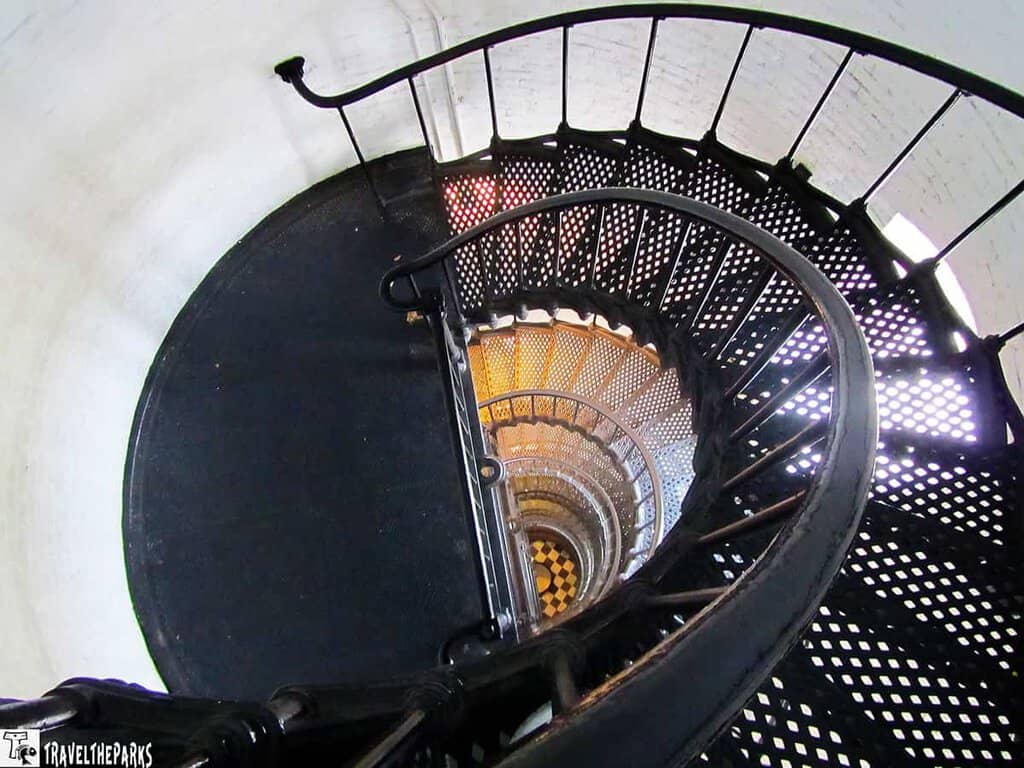 a spiraling metal staircase inside St Augustine lighthouse, viewed from above. The spiral design creates concentric circles that draw the eye towards the center of the image. The stairs are made of dark metal with perforated steps and are bordered by a sturdy handrail. The central column of the staircase is visible, with light filtering through the perforations, casting intricate patterns. The surrounding walls are a smooth, light-colored masonry, adding contrast to the dark staircase. At the bottom, a black and white checkered floor is faintly visible.