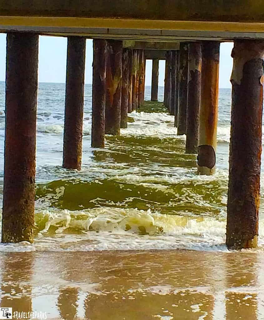 A view underneath the St Augustine pier extending into the ocean. The photograph is taken from the shore, capturing the alignment of weathered, rust-colored metal columns supporting the pier. These columns diminish in perspective towards the center, creating a tunnel-like effect. The ocean appears calm, with gentle waves lapping against the columns and reaching the sandy beach. The sky is visible in the distance, adding a light backdrop to the scene.