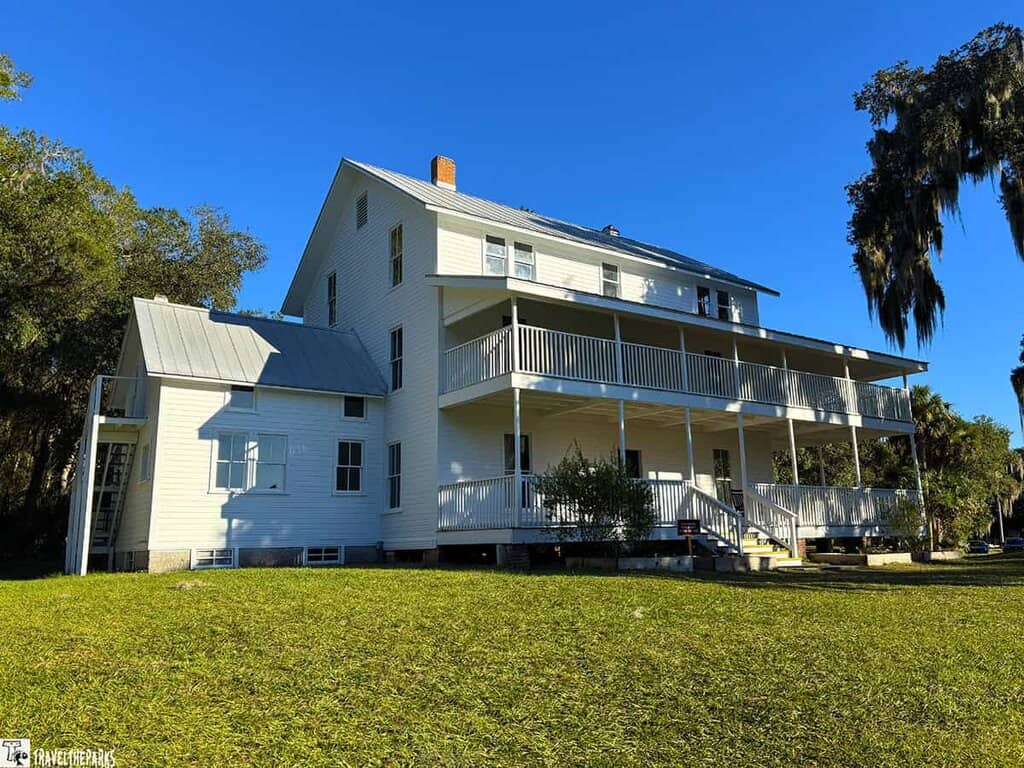 A historic white house with a two-story porch and metal roof at Blue Springs State Park, surrounded by greenery and trees under a blue sky.