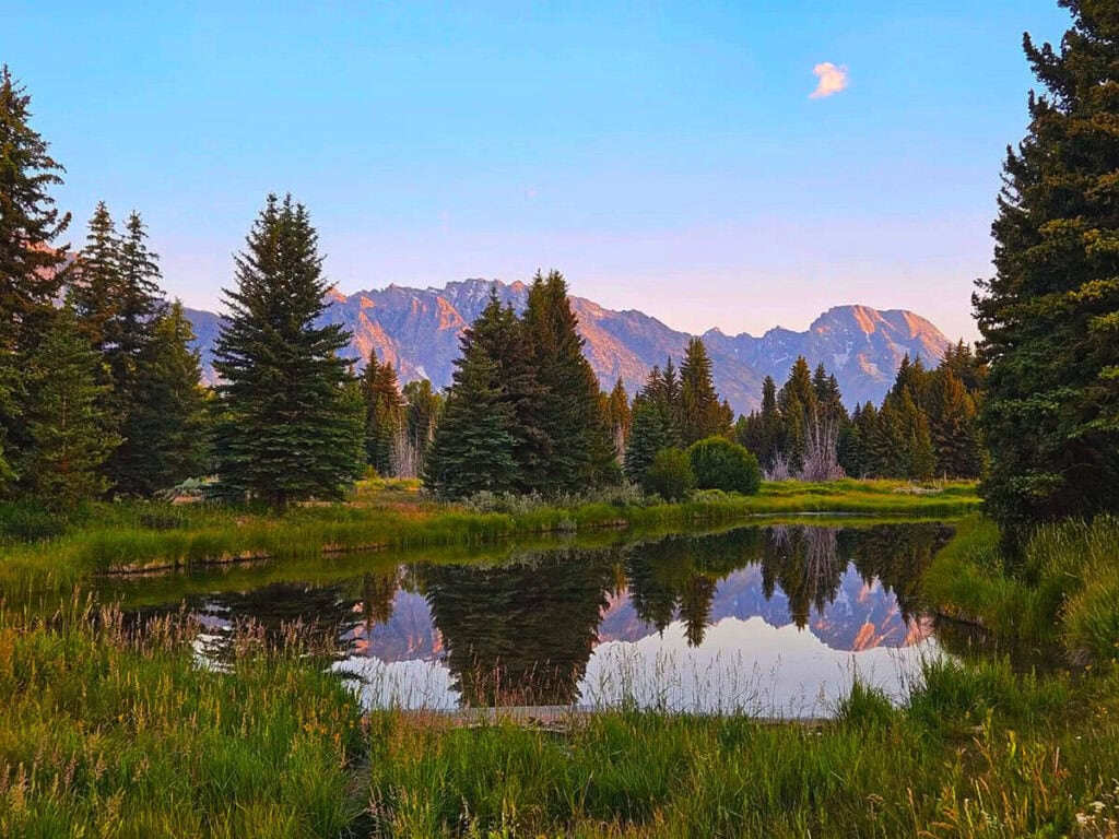A reflective body of water with surrounding trees and distant mountains under a blue sky in Jackson Wyoming