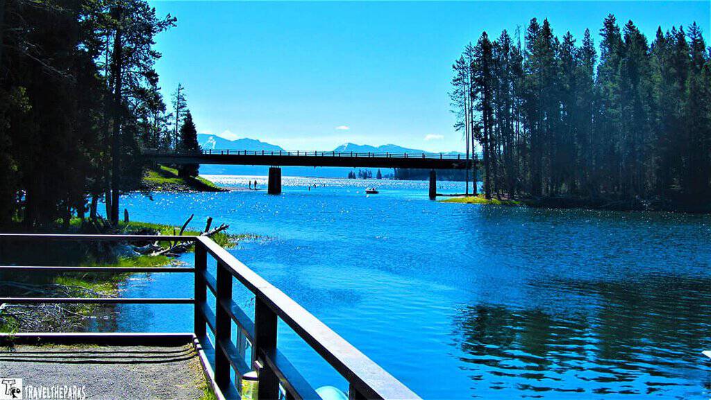 Bay Bridge Marina, Yellowstone Lake-A lakeside view with a wooden bridge over the water, pine trees on both sides, and mountains in the background.