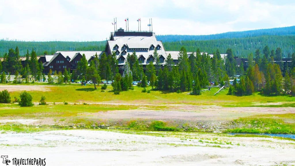 Old Faithful Inn-Yellowstone National Park: A large, rustic building with flags on the roof, surrounded by evergreen trees and grassy fields.