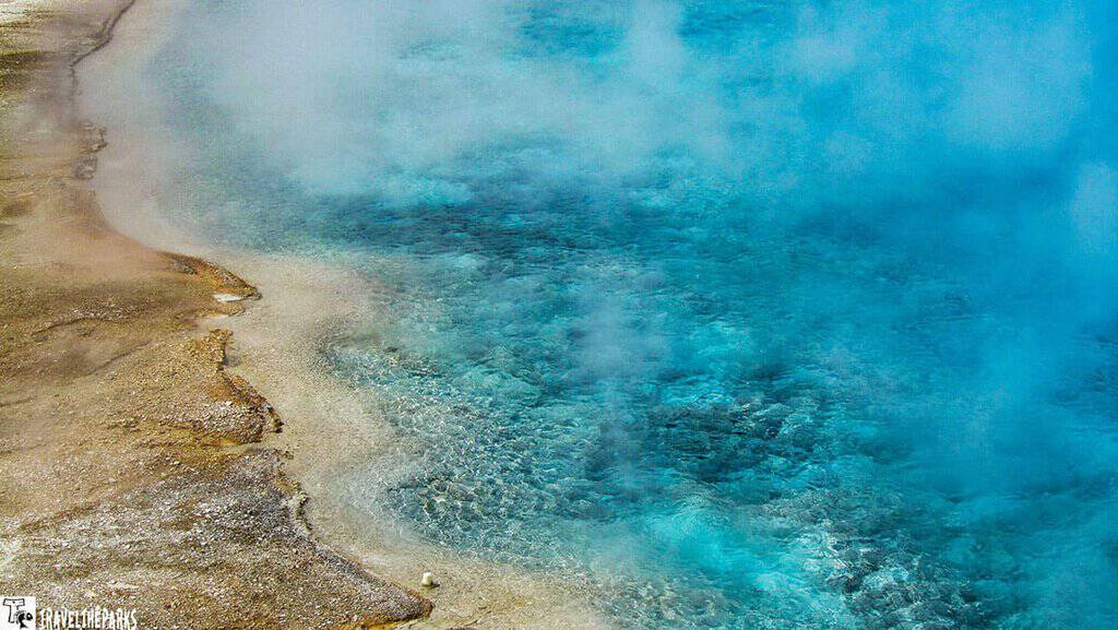 Excelsior Geyser: A vibrant blue hot spring with steam rising, surrounded by rocky terrain. A wooden railing is visible in the background, evoking a serene, natural wonder.