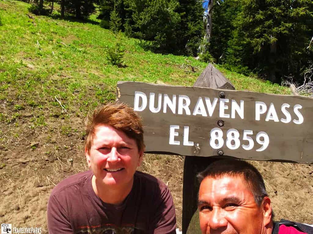 Two people smiling in front of a sign reading "Dunraven Pass EL 8859" with a grassy and wooded background.

