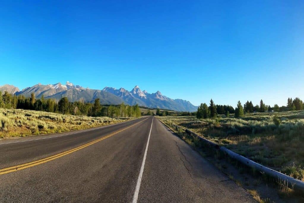 A road leads to the Grand Teton mountains under a clear blue sky.

