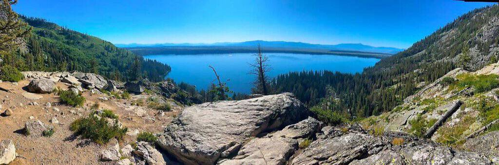 Panoramic view of Jenny lake surrounded by forests and mountains under a clear blue sky. Taken from Inspiration Point Overlook.