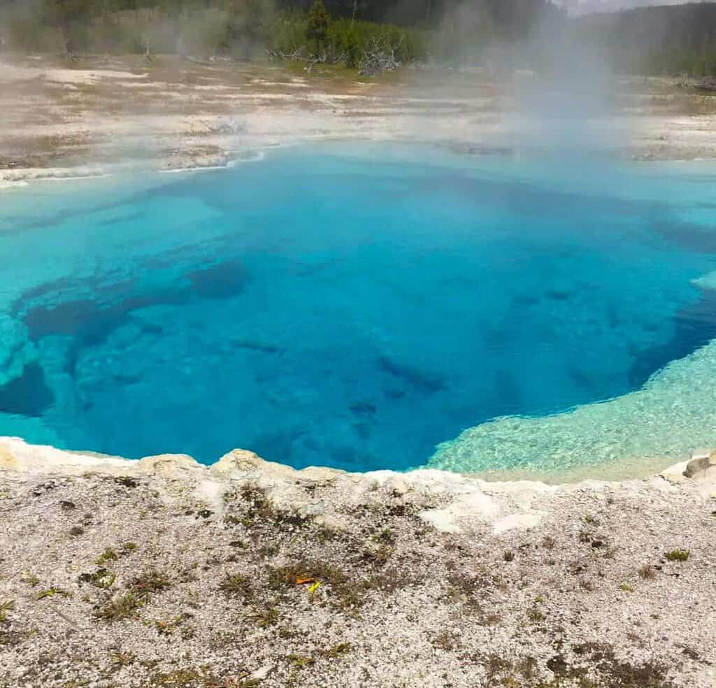 sapphire-pool geyser: Steaming blue hot spring with vivid, clear water against a vast, open landscape. The scene is serene, with a bright sky and distant surrounding trees.
