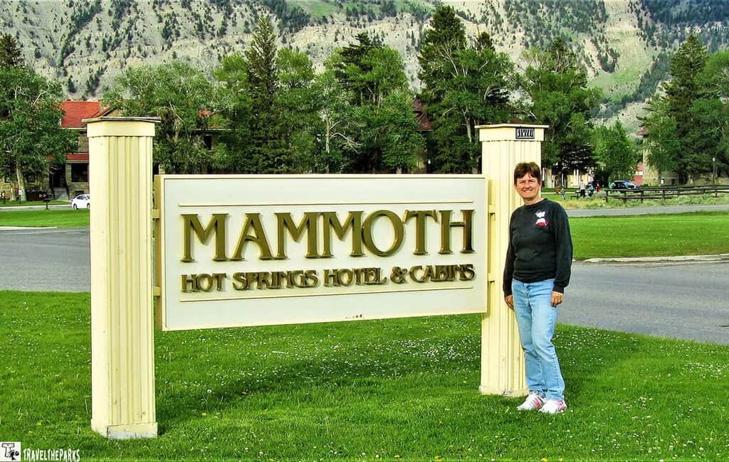 A woman poses in front of a sign that reads "Mammoth Springs," with a scenic background behind her at Yellowstone National Park.