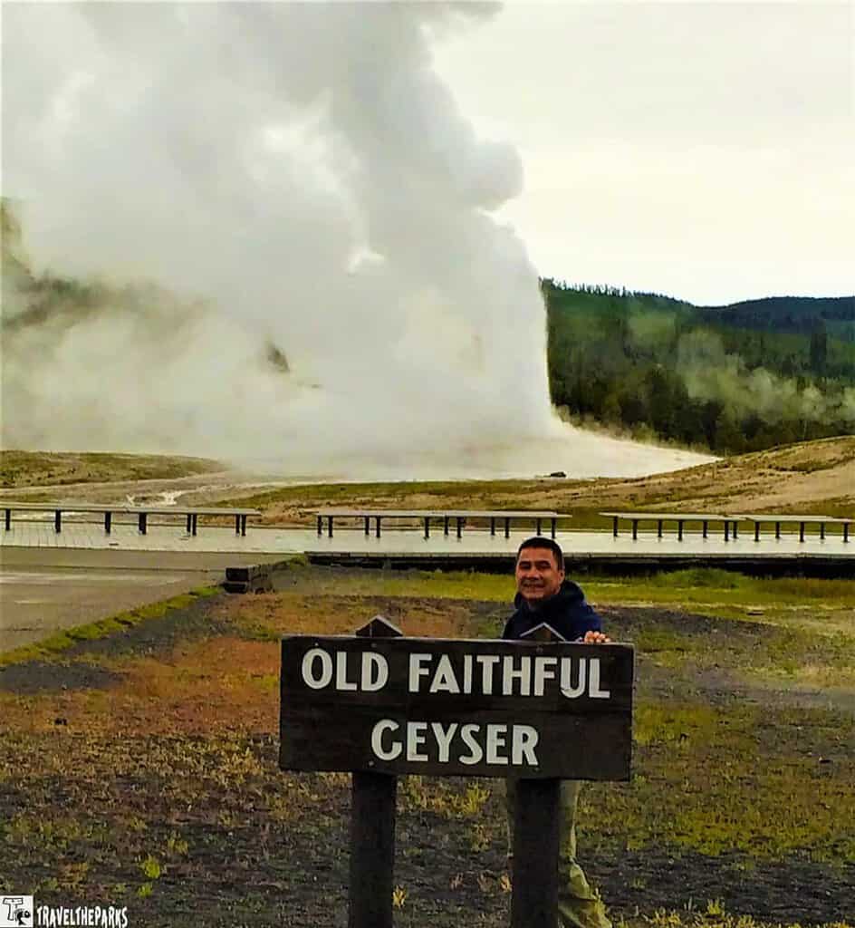 Person smiling behind an "Old Faithful Geyser" sign at Yellowstone. The geyser erupts in the background, surrounded by a cloudy sky and greenery.