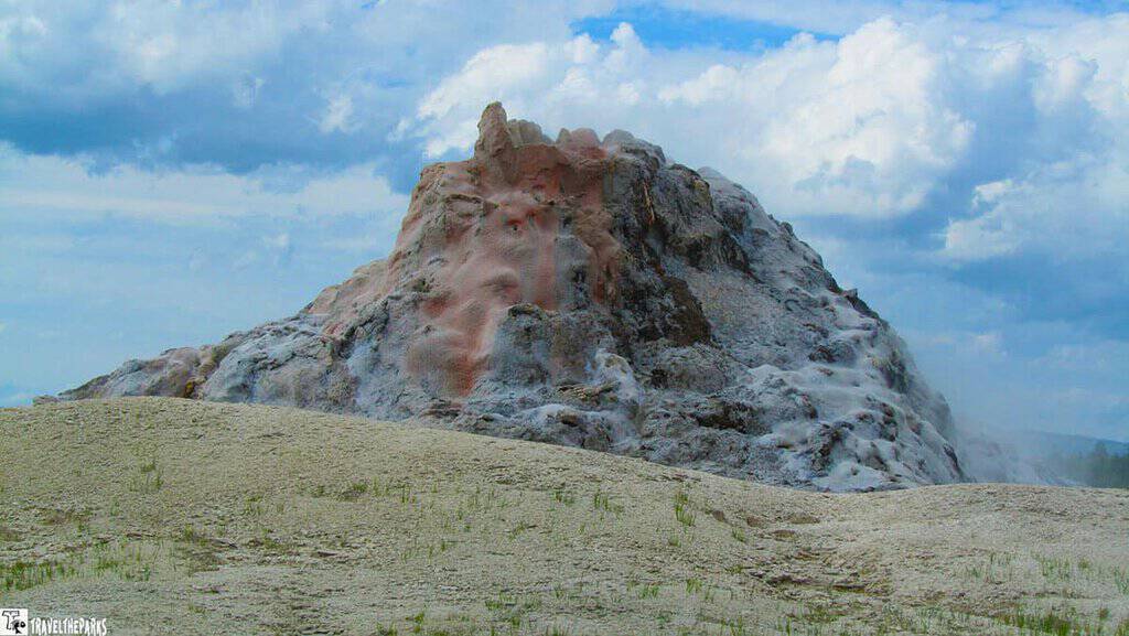 White Dome Geyser erupting from its mineral-covered cone in the Lower Geyser Basin of Yellowstone National Park, sending a plume of steam and water into the air against a forested backdrop.
