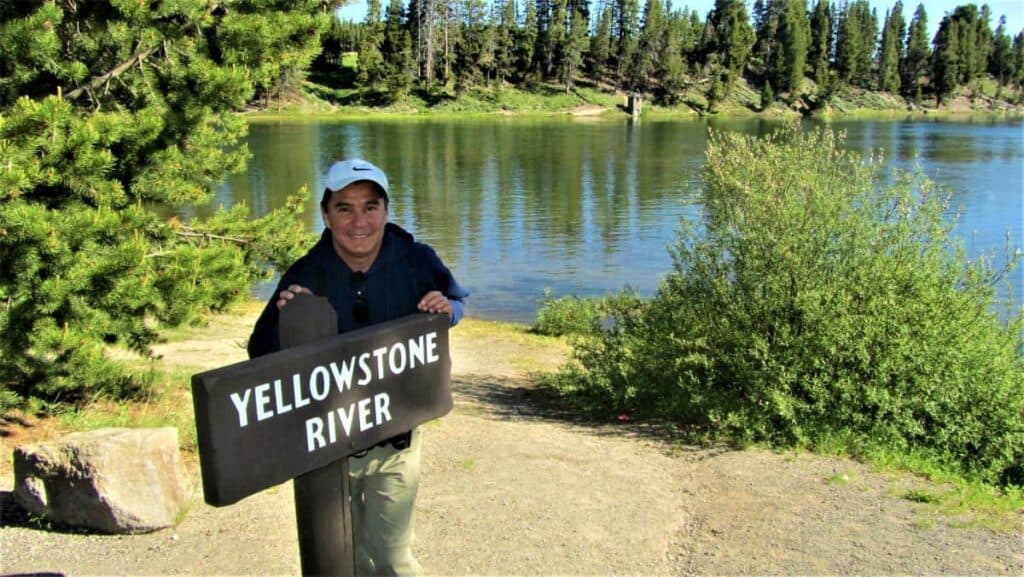 Man stands beside a "Yellowstone River" sign, smiling against a scenic backdrop of calm water, lush greenery, and pine trees under a clear sky.