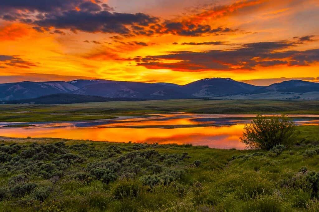A stunning sunset over Lamar Valley in Yellowstone, with a colorful sky, rolling mountains, and a reflective body of water surrounded by green vegetation.