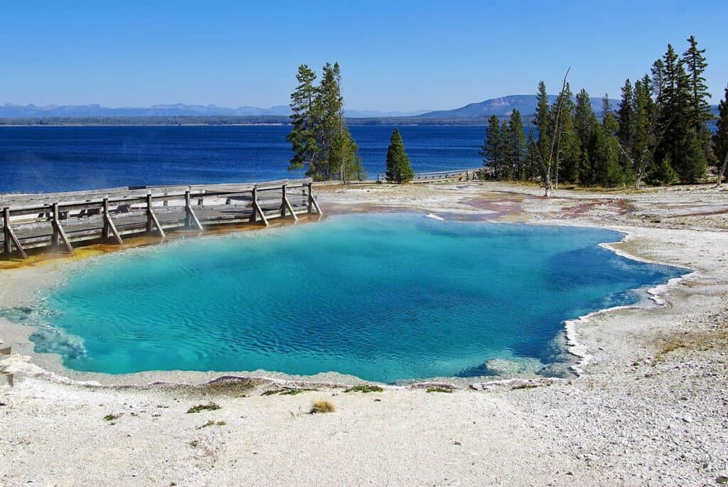 Black Pool - West Thumb Geyser Basin-A colorful geothermal hot spring near a blue lake and forested area.