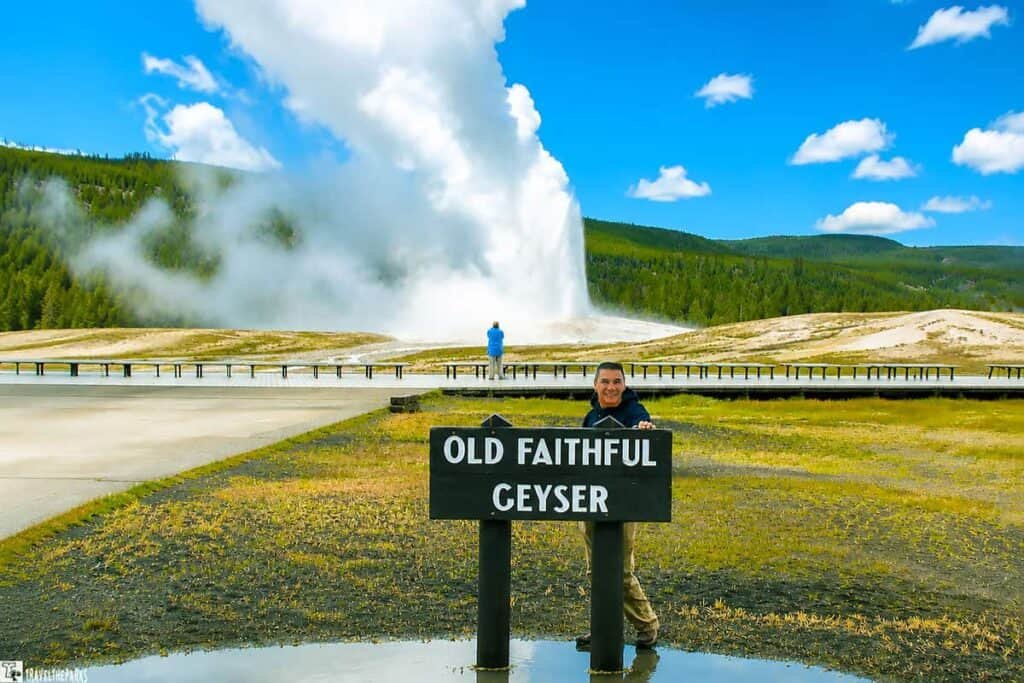 Person smiling behind an "Old Faithful Geyser" sign at Yellowstone. The geyser erupts in the background, surrounded by a cloudy sky and greenery.