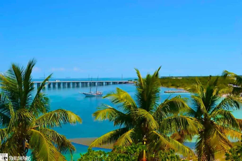 A tropical coastline with palm trees, sailboats, and a bridge over azure water under a blue sky.