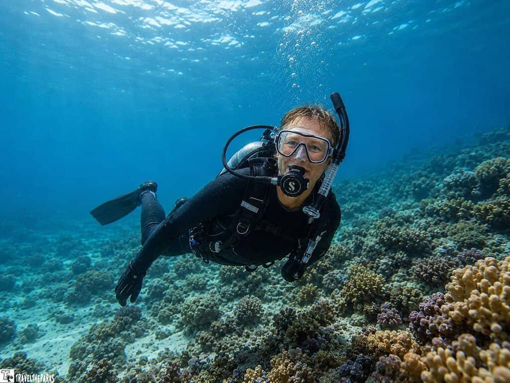 A scuba diver with a mask and fins swims over a coral reef in clear blue water, with sunlight filtering through the surface.