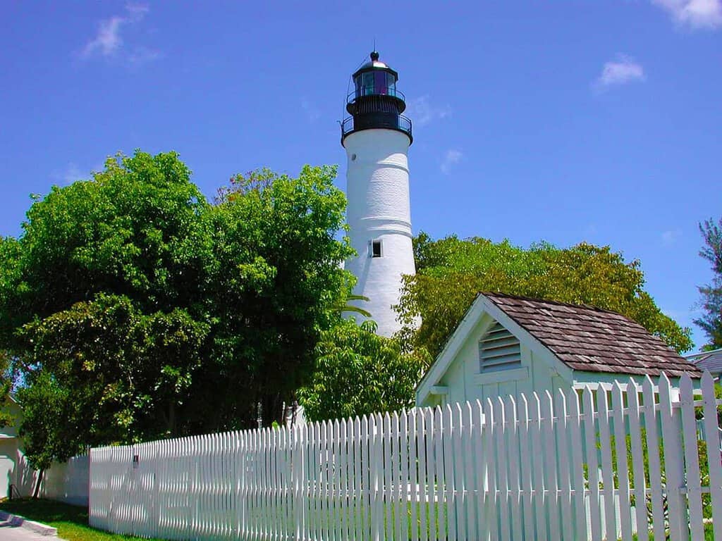 Tall white key West lighthouse with a black top, surrounded by green trees against a blue sky.
