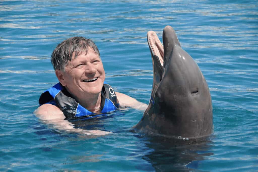 A person smiling in the water with a dolphin beside them wearing a blue life jacket.