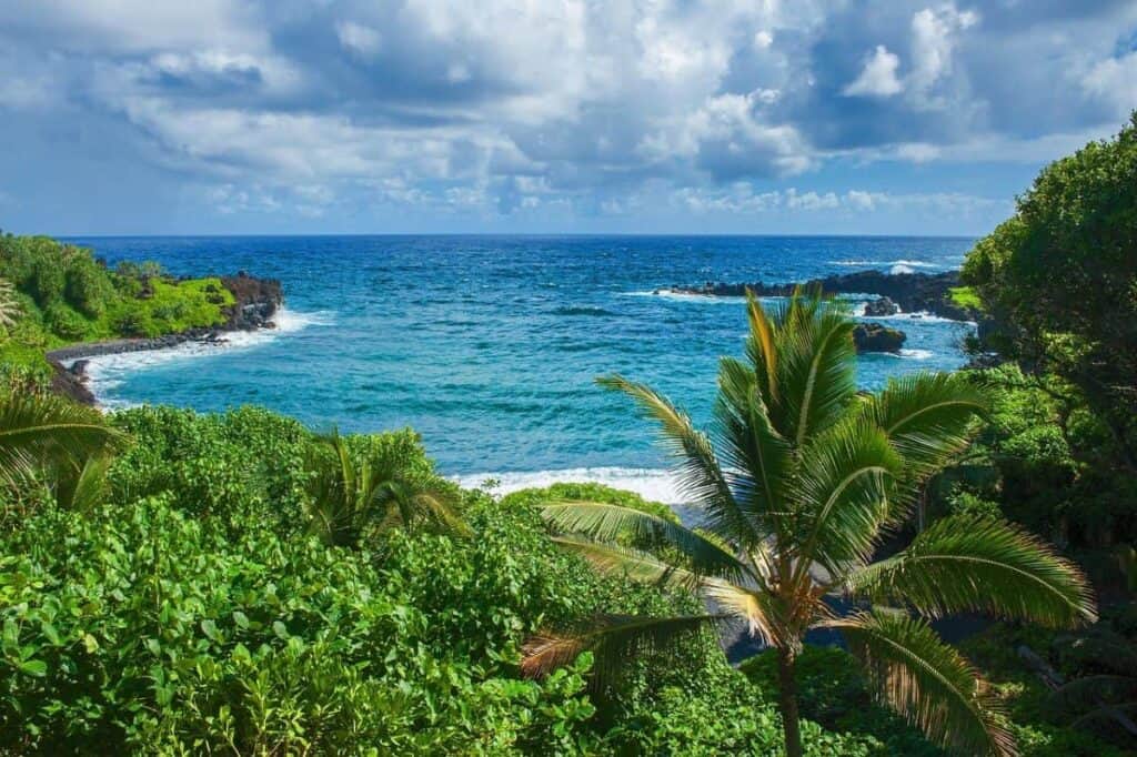 Tropical coastline on the drive to Hana with lush greenery, rocky shoreline, and a blue ocean under a partly cloudy sky.