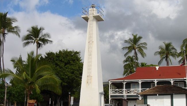 Lahaina Lighthouse surrounded by palm trees and a red-roofed building against a partly cloudy sky.
