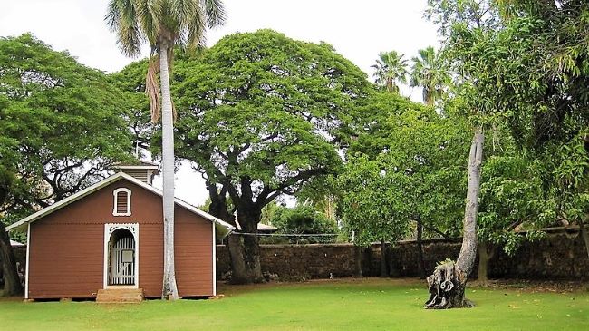 Hale Pa'ahao - Old Lahaina Prison: A small brown building with white trim on a green lawn, surrounded by large trees and a palm tree.