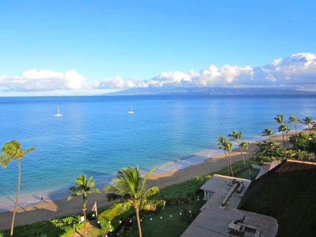 View of Kaanapali beach in Maui with palm trees and sailboats, with an island and clouds in the distance.