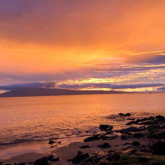 A vibrant Maui sunset over an ocean with a rocky shoreline in the foreground.