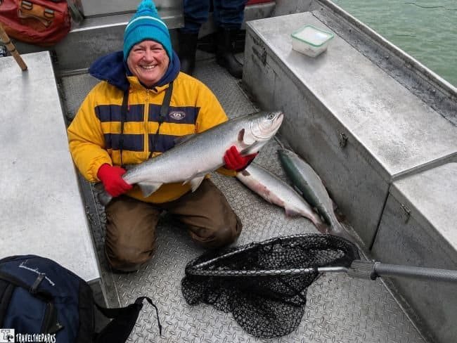 Smiling person in a yellow-and-navy jacket kneeling in a fishing boat and holding a large silvery fish, with two other fish and a landing net nearby.

