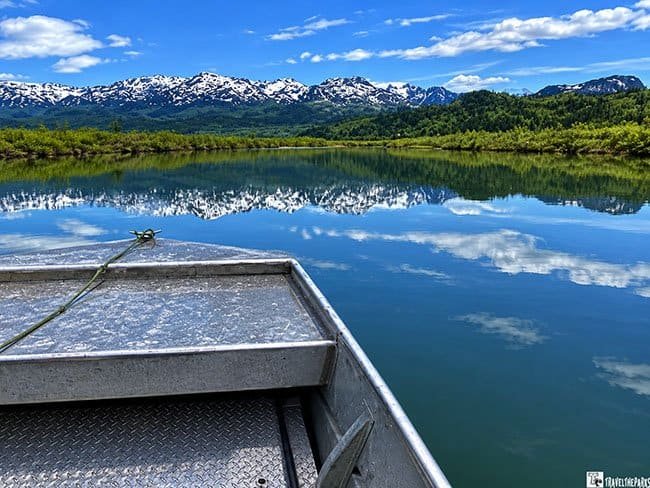 A metal boat on a calm lake with snow-capped mountains in the background.