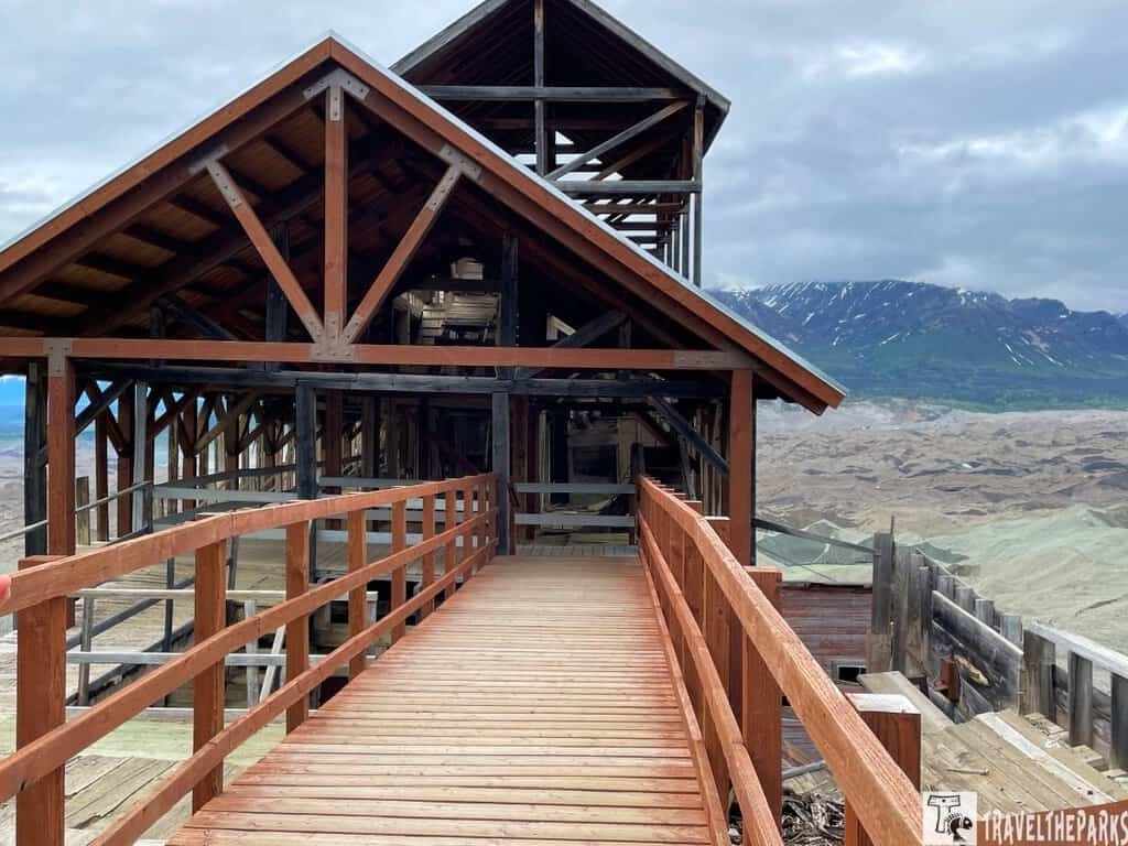 A wooden walkway leads into the Kennecott Mines National Historic Landmark, surrounded by mountainous terrain and an overcast sky.