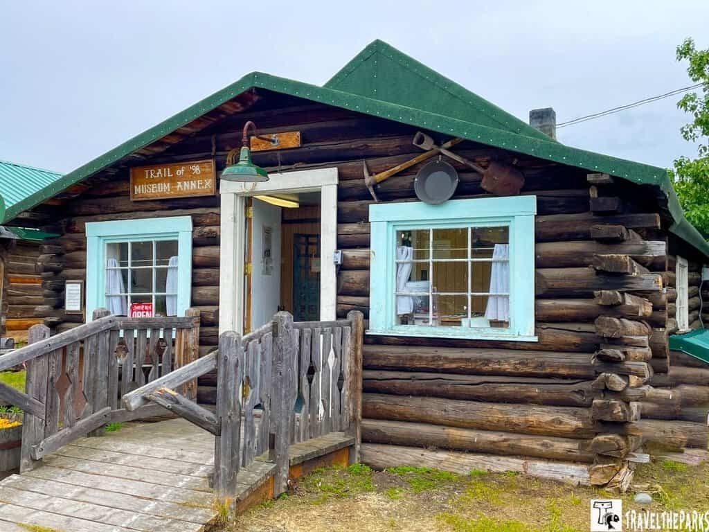 A rustic log cabin with a green roof and pale blue window frames, featuring a sign for the "Trail of '98 Museum - Annex."