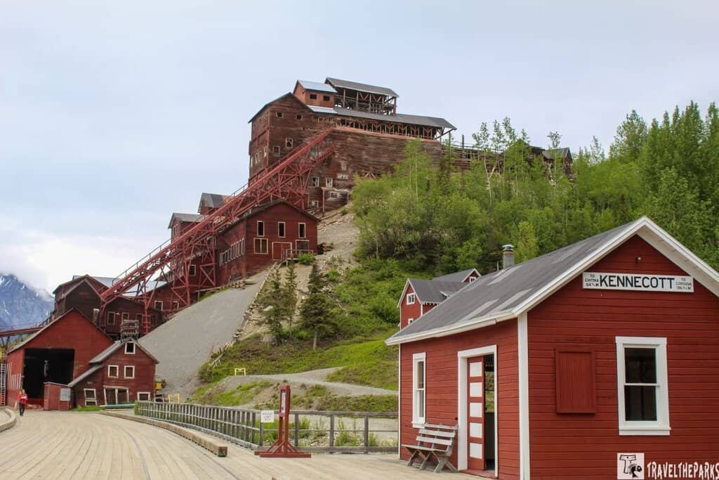 A series of historic red mining buildings at Kennecott Mines National Historic Landmark, with a conveyor belt leading up the hillside and a wooden walkway in the foreground.